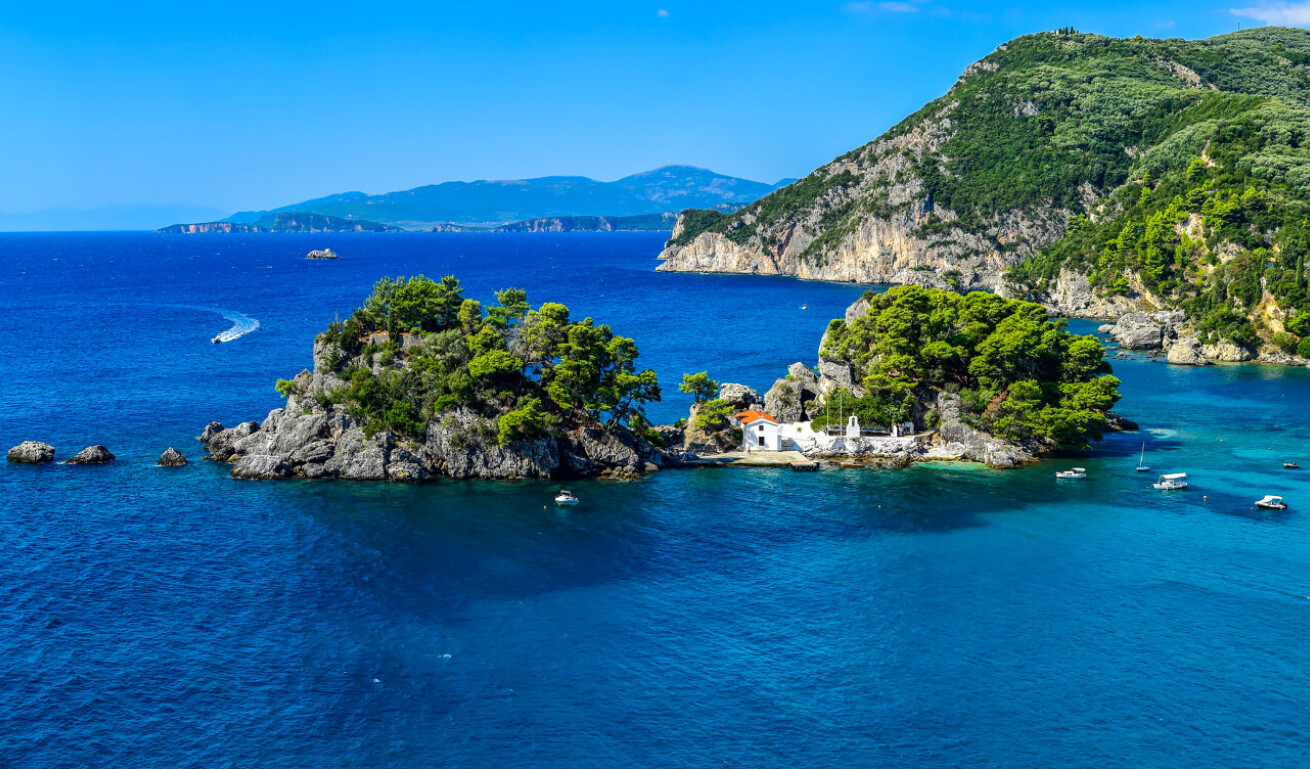 View of the port of the town with blue waters, a rocky islet with a chapel in the background and sailing boats