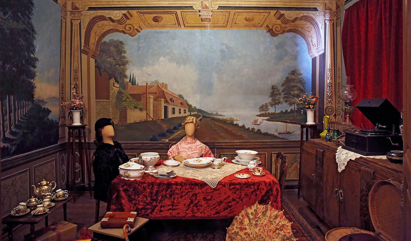Folklore museum interior room with painted walls. To the right red curtain and wooden furniture with gramophone on top. In the middle of the room a table with a red tablecloth, white tableware with paintings. 2 faceless dolls in traditional costumes sit on chairs.
