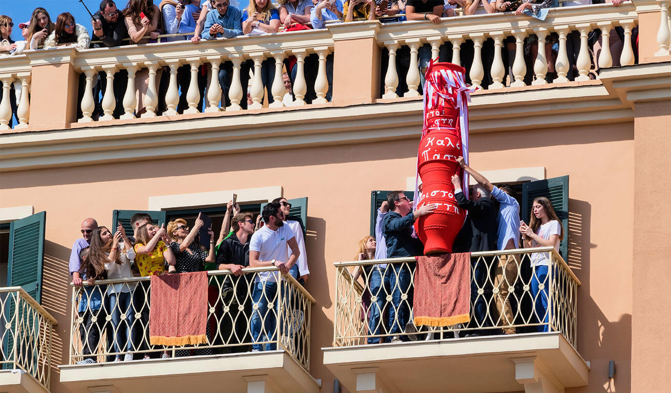 Facade of a building with balconies full of people. On one balcony they are holding a large red jug at the edge. Sunshine.