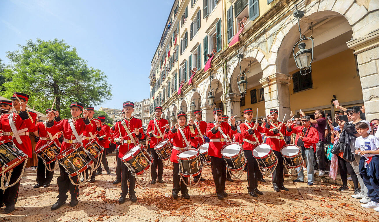 Philharmonic orchestra with drums and red jackets in front of a traditional building with arches. Sunshine.
