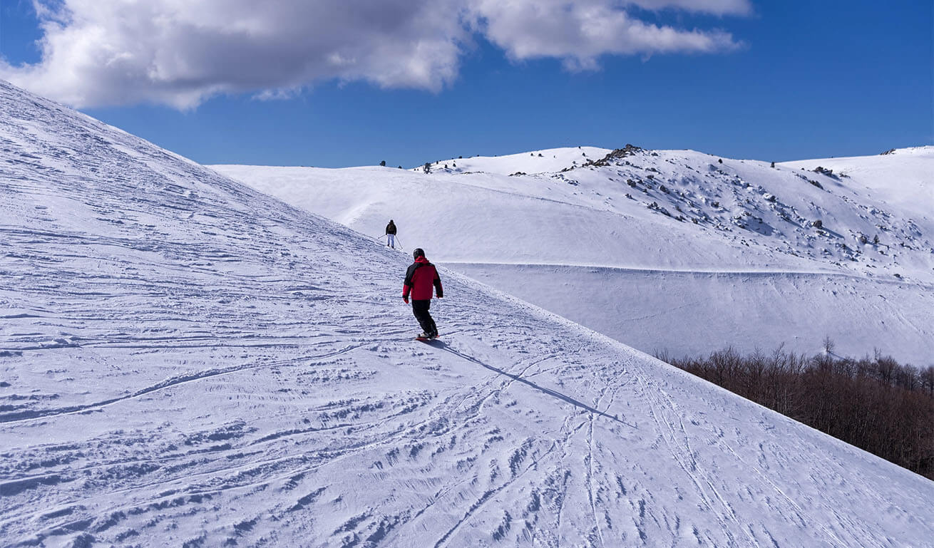 Snowy ski slope with ski tracks. Two people snowboarding and skiing. Sunny with a few clouds.