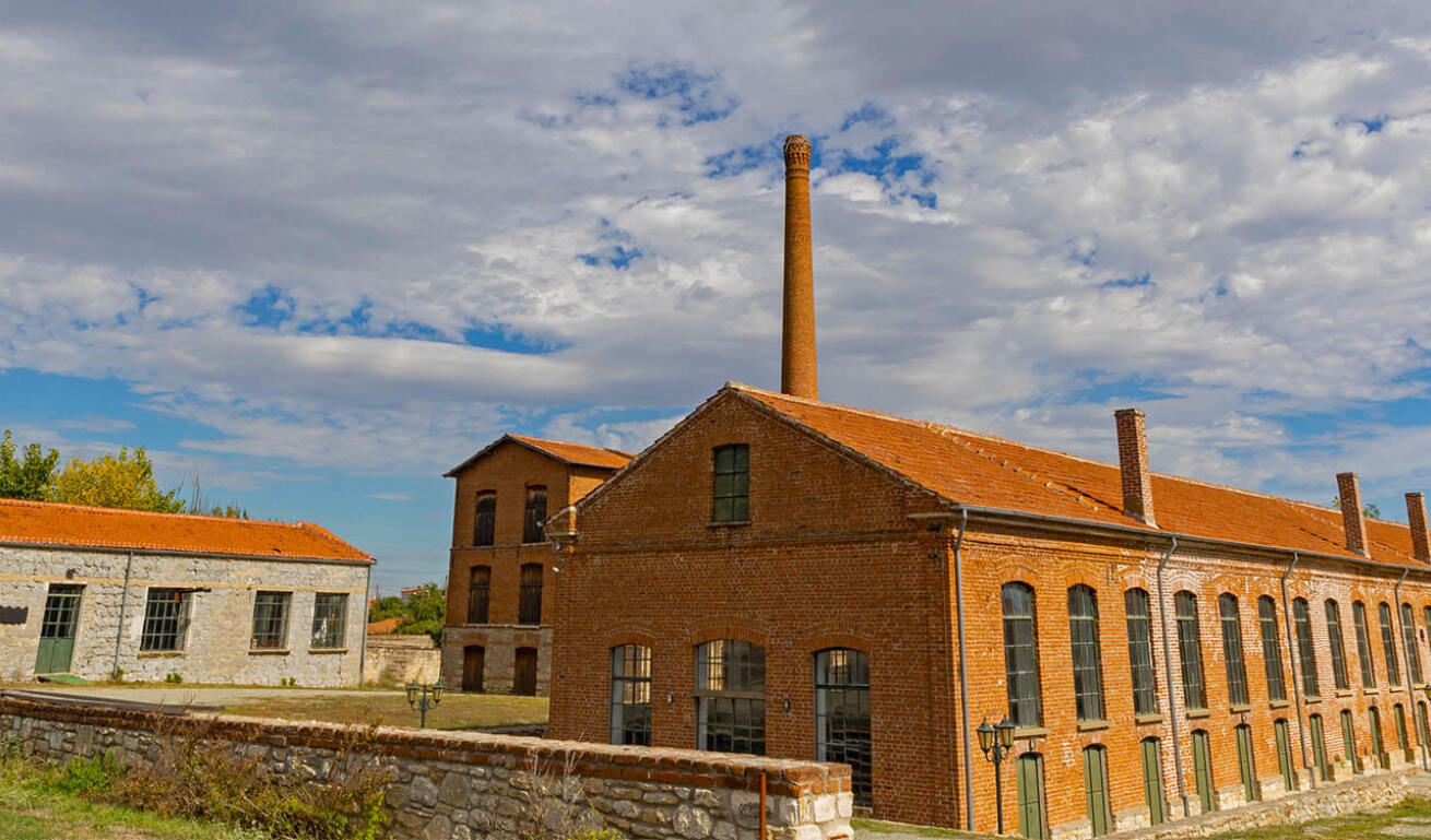 Historical industrial building with brick walls, tall windows, and a chimney. The surrounding area is open, with stone buildings and greenery under a partly cloudy sky. 