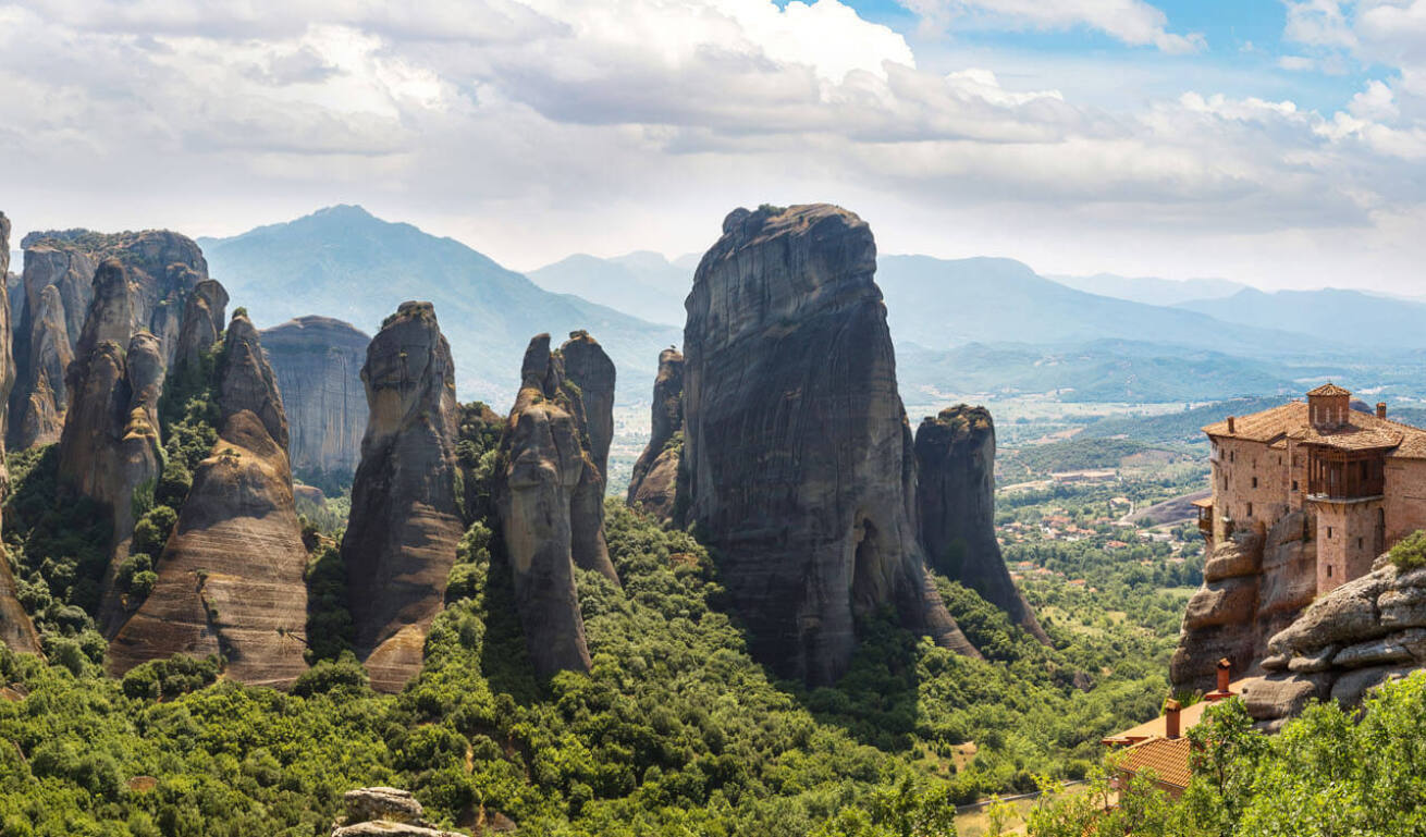 A panoramic view of high cliffs over a green valley. In the foreground there is a traditional stone monastery.