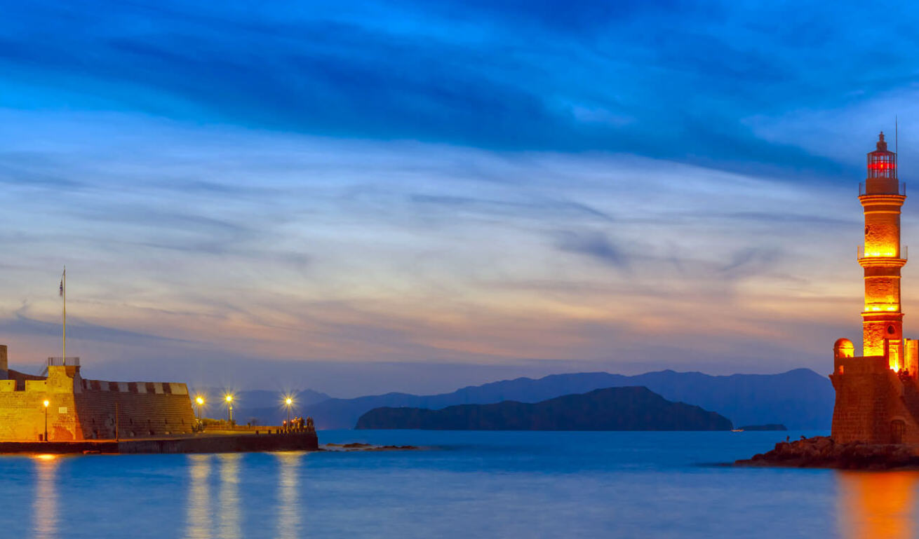 Είσοδος λιμανιού με χαμηλά τείχη από την μια πλευρά και πέτρινο φάρο από την άλλη με αναμένα φώτα το δειλινό.