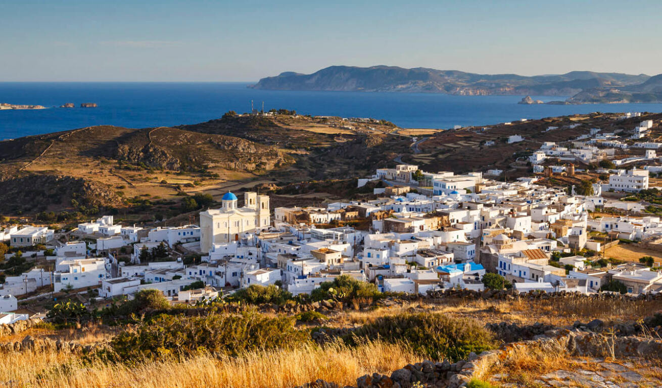 View from above of a settlement with white houses and a large church. Around low hills with sheaves. In the background sea and island. 