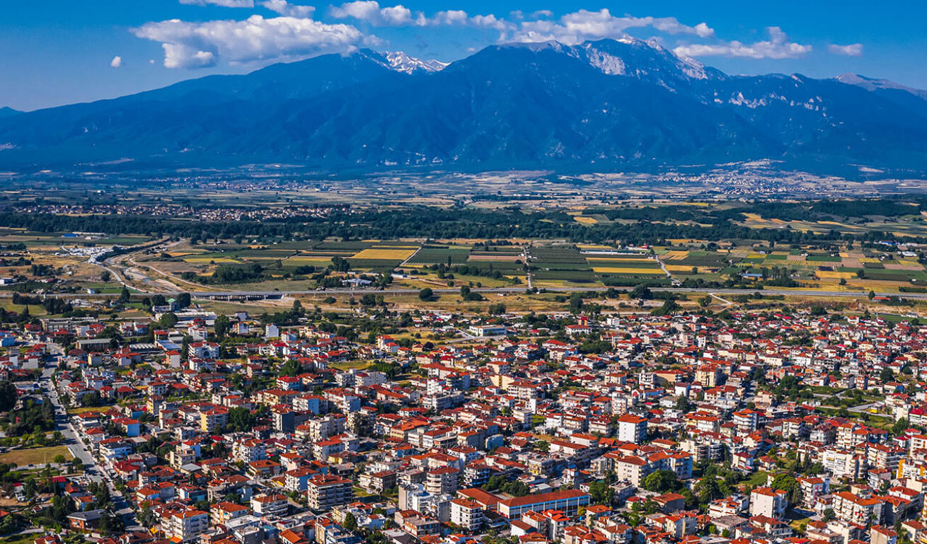 Aerial view of city with low buildings with red roofs. In the background fields and in the distance high mountains. Sunshine.