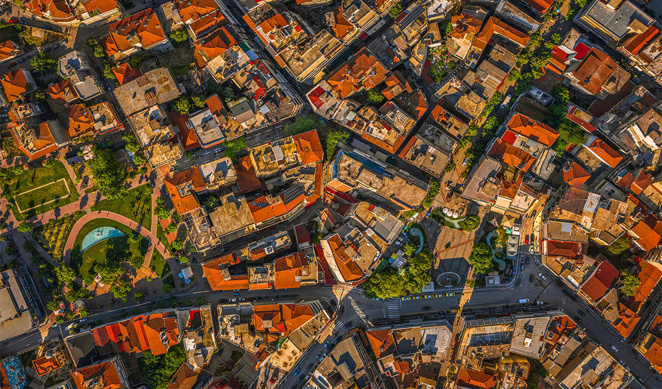 Aerial view of a dense urban area with red-roofed buildings, winding streets, and green spaces in a lively cityscape.