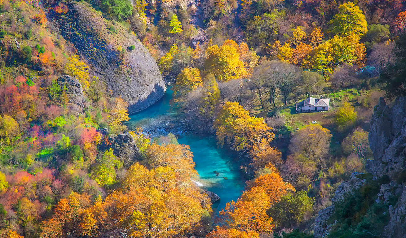 Blue water river running through lush forest with autumn colors at the base of a canyon. There is a building with a sloping stone roof.