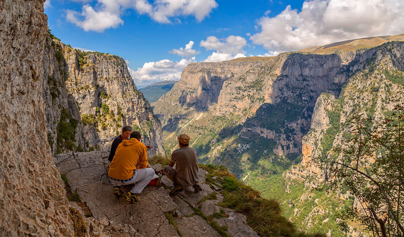 Three people sitting on a rocky ledge overlooking a deep mountain gorge under a bright sky.