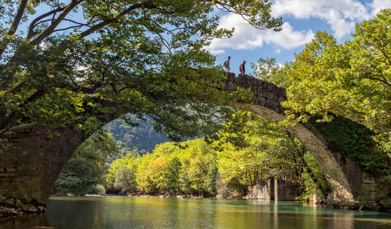 Stone bridge with an arch over a calm river with dense vegetation. Two people are walking on it. 