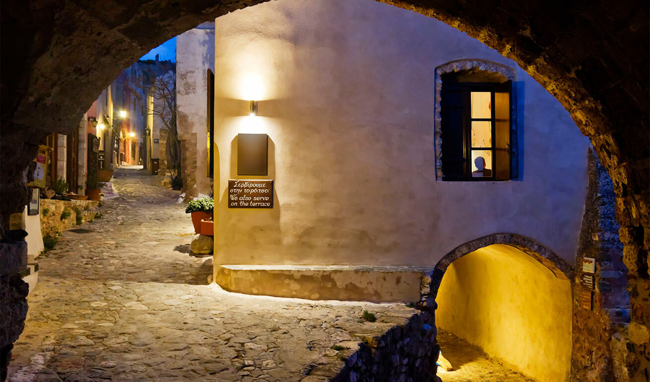 A charming cobblestone alley in Monemvasia, Greece, lit by warm lights, with an arched passage and a cozy window view at night.