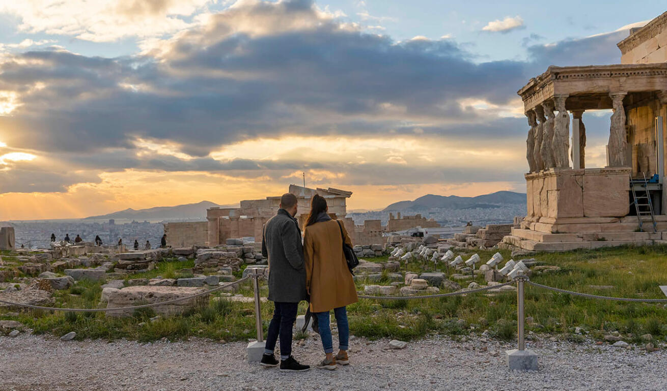 Couple at an archaeological site. Ancient marble buildings with statues, columns and pediment. View of the city and sea. Cloudy with sun.