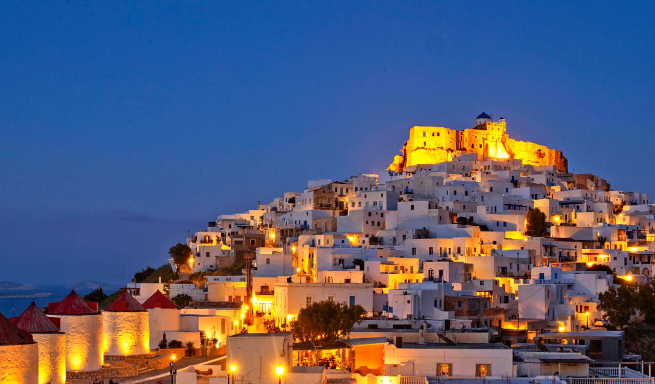 Night shot with lit lights of a settlement with small white houses on a hill with a castle and a church at the top. 