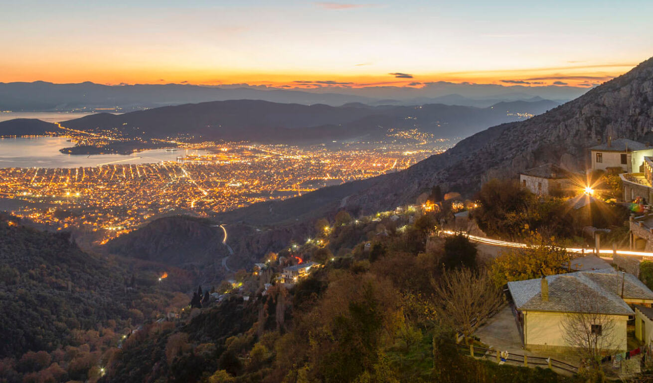 View from above of a seaside town at sunset with lights on. In the foreground traditional houses on the mountain slope. 