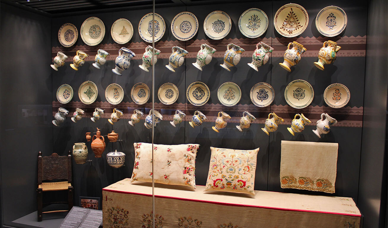 Large glass museum display case with decorative plates, jugs, fabrics and other objects.
