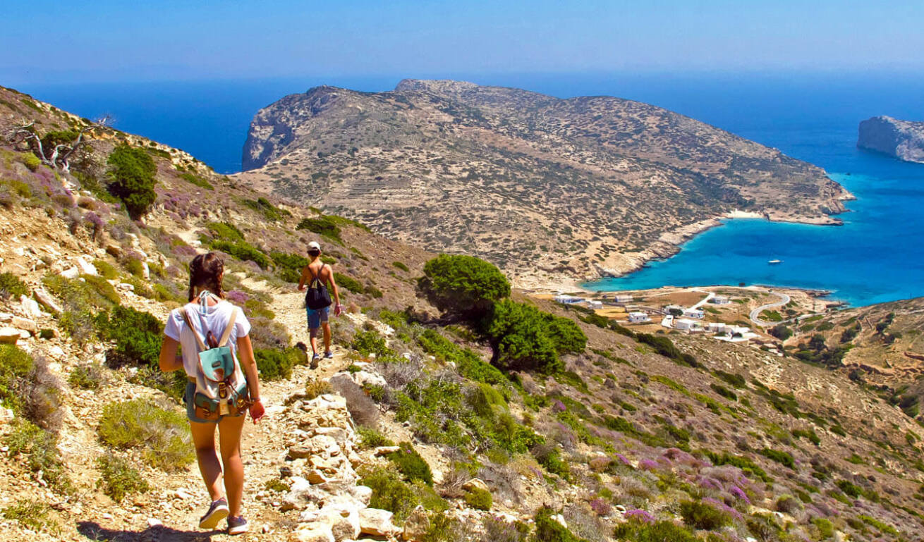 2 young people walking on a path with low vegetation. In the distance is the sea, a few white houses and a small island. 