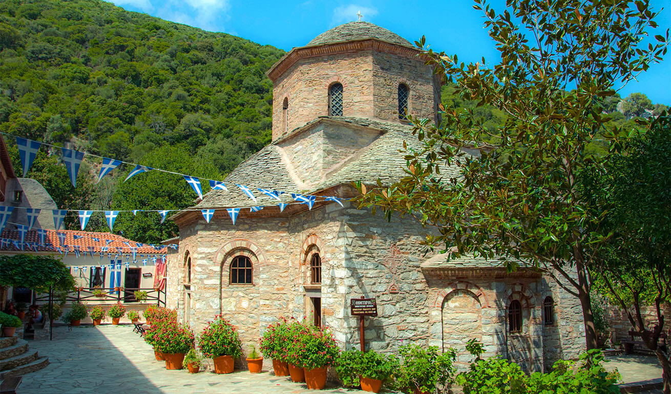 Traditional stone church with a dome, decorated with flags, in a paved courtyard with small flower pots. Surrounded by a lush green hillside. Sunshine.
