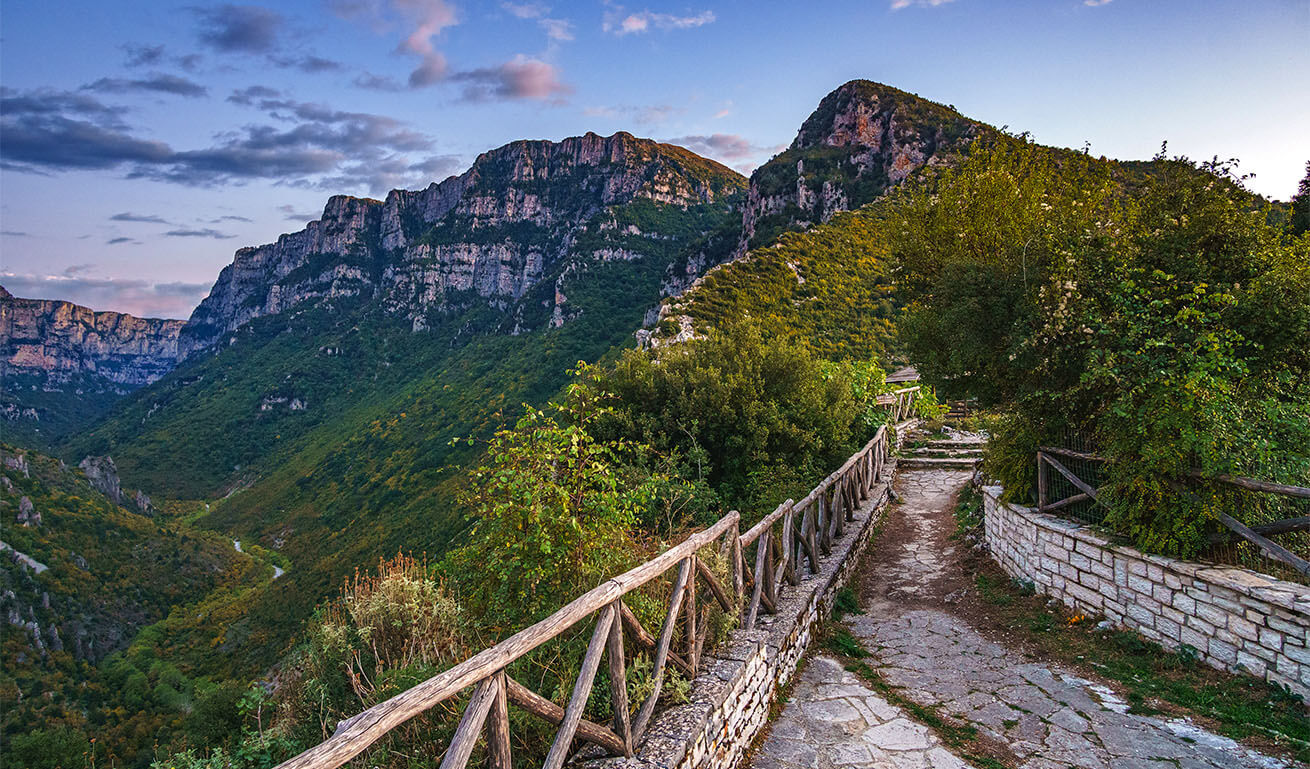 Paved path with a wooden fence and a view of the canyon. Green steep slopes and high cliffs. Clear sky with few clouds in the evening.