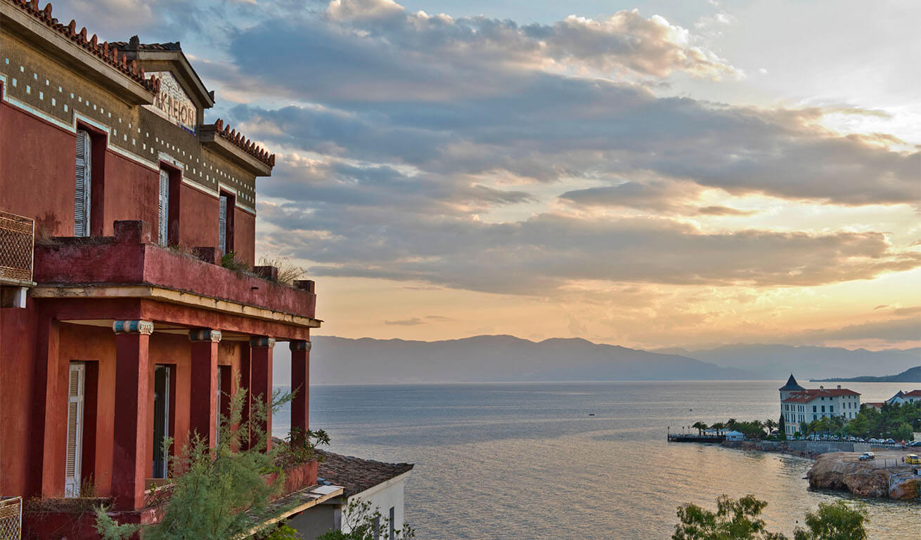 On the left, an old red building with a balcony, cornice and columns with white Ionic capitals overlooking the sea and a settlement beach. Evening with few clouds.