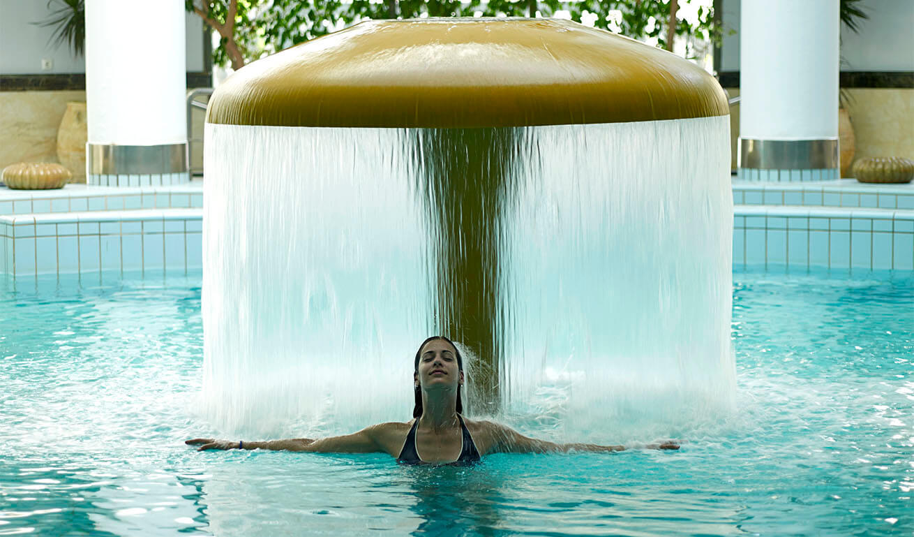 Swimming pool with a circular fountain that flows water. A girl in a swimsuit is inside and enjoying herself. Around you can see 2 white columns and plants.