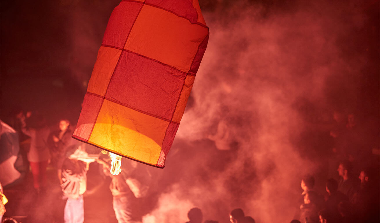 A small reddish lantern with a flame rises towards the sky around a crowd of people. Night with smoke.