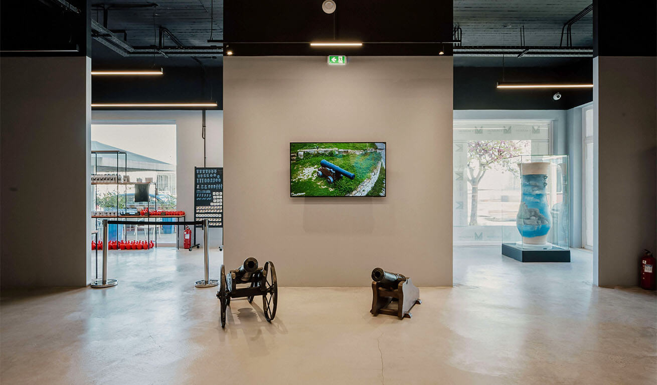 Interior of a museum with 2 cannons and a picture of a cannon and 2 openings showing a large blue painted jug and a display of small red jugs.