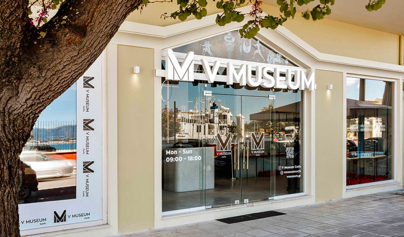 Glass museum facade with the museum's name, framed by a light-colored wall. Reflecting low buildings and a clear sky.