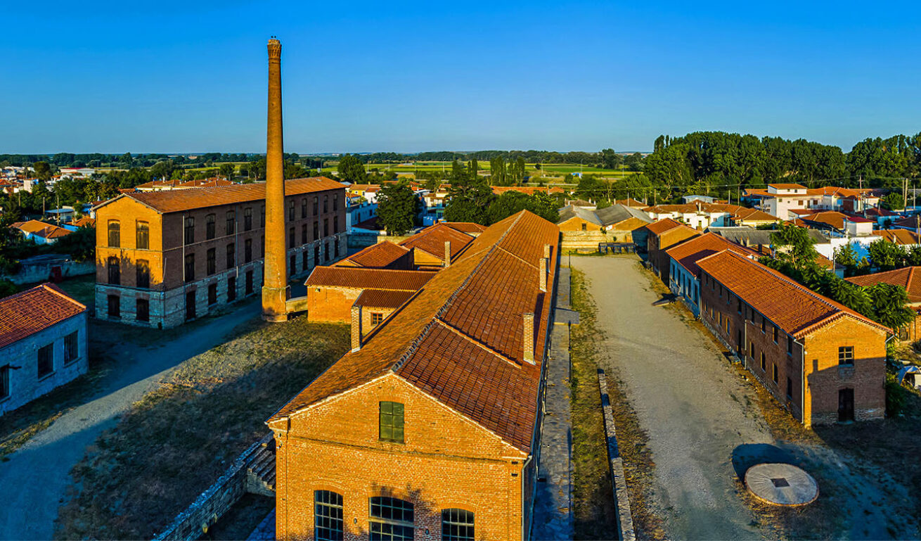 Historic stone industrial buildings with tiled roofs and a chimney. In the background, a traditional settlement on a plain with trees. Sunshine.