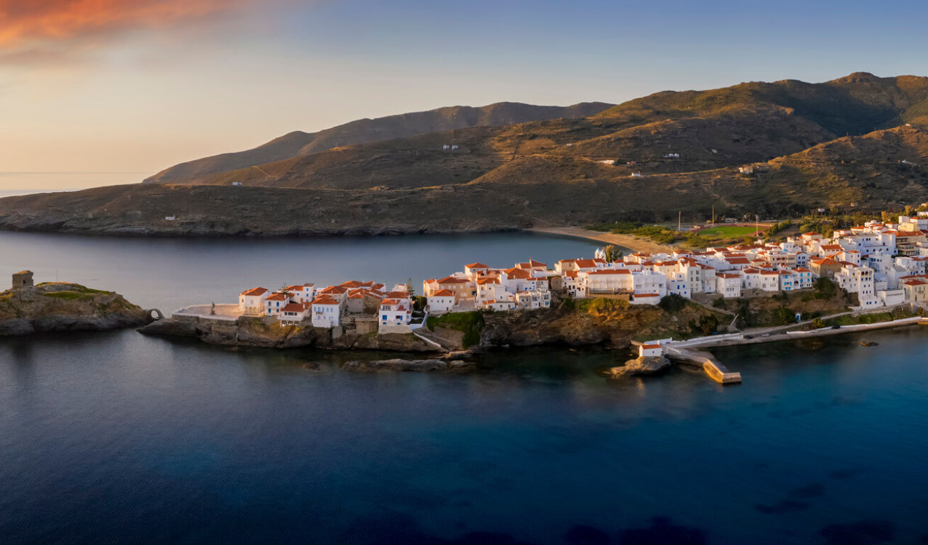 Aerial view of a small peninsula with a traditional Cycladic settlement at sunset. In the background you can see a beach and hills. 
