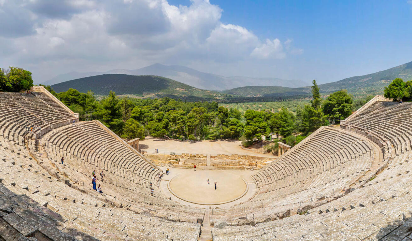 View from above of a large ancient theater and in the background a green valley with distant hills. 