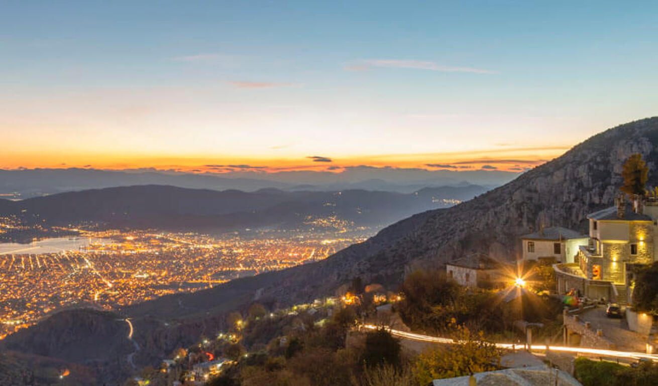View from above of a seaside town at sunset with lights on. In the foreground traditional houses on the mountain slope. 