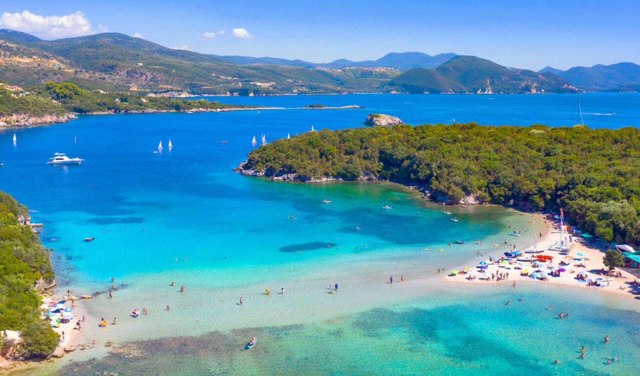 Παραλία με πεύκα και γαλαζοπράσινα νερά, με πολλούς λουόμενους και ιστιοπλοϊκά