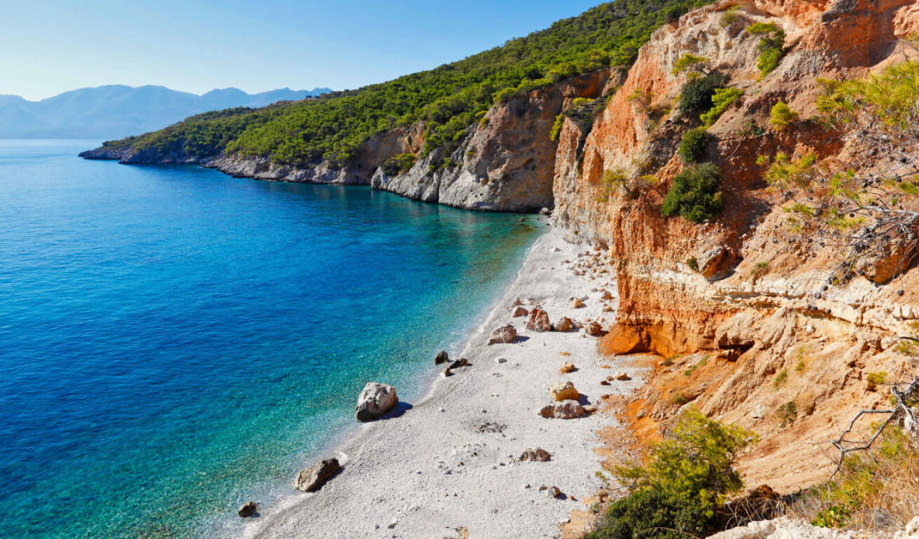 A beach with greenish blue waters, redish rocks and pine trees
