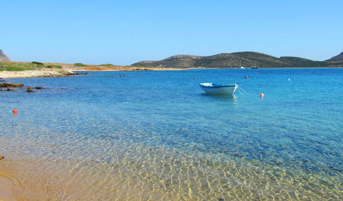 Small boat in front of a beach with sand and crystal clear waters and in the background small hills. 