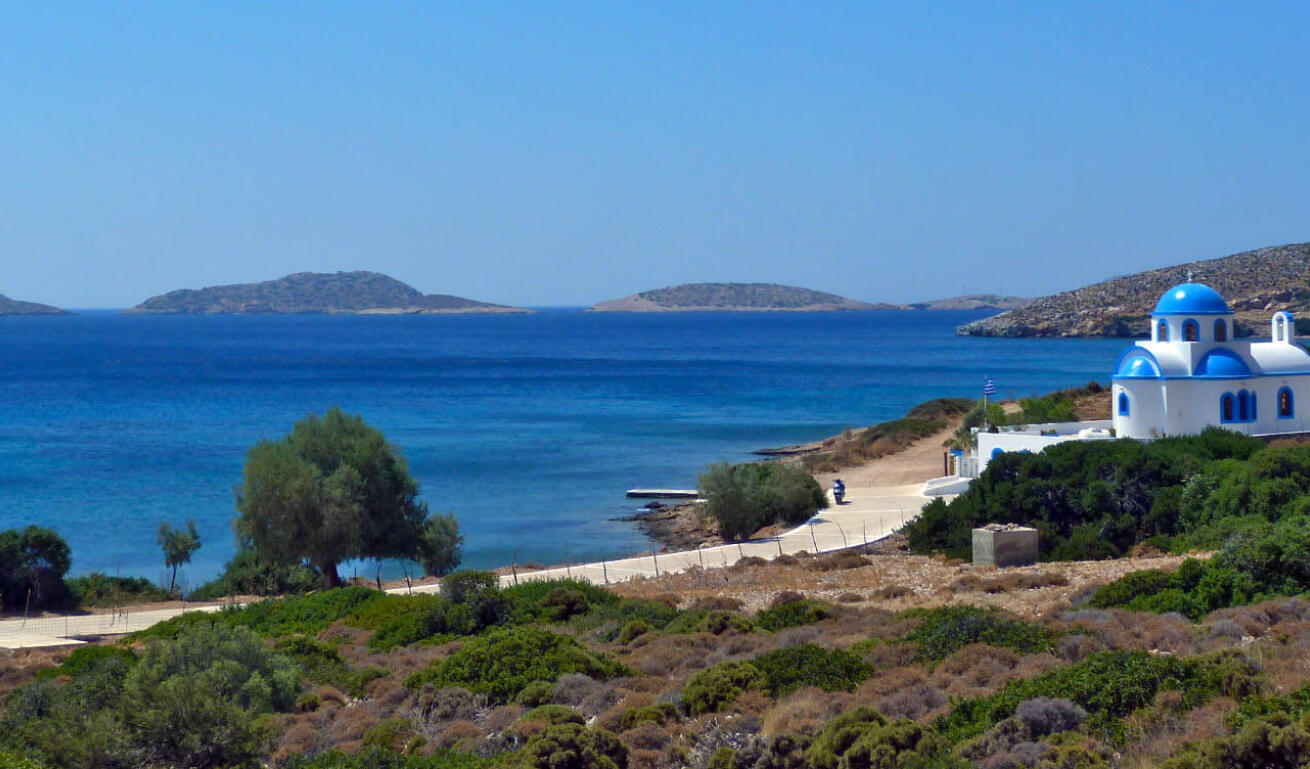 Παραλία του νησιού με πεύκα και θέα τη γαλάζια θάλασσα και στο βάθος μία εκκλησία με μπλε τρούλο