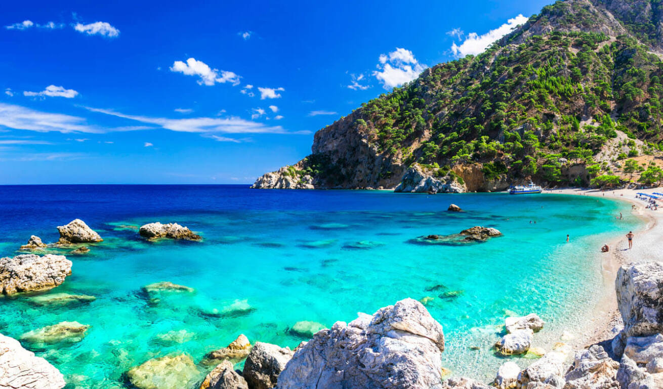 Beach with greenish blue waters, rocks and pine trees as well as swimmers walking around