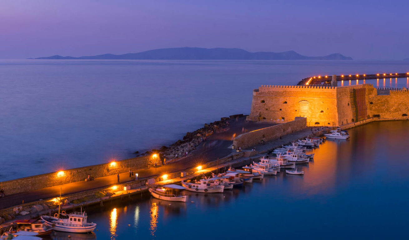 Pier with boats and a small fortress made of stones at sunset with the lights on.