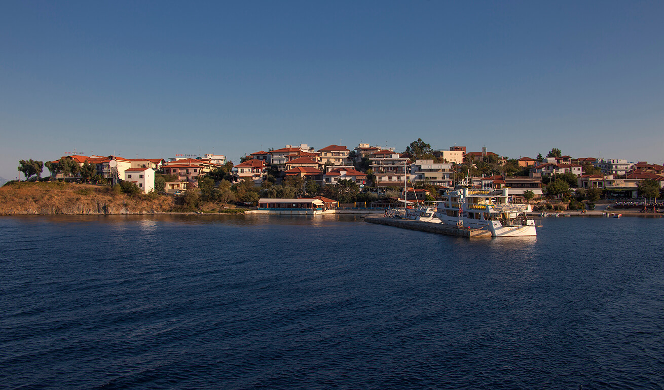 A panoramic view of a coastal town at sunset with buildings along the shore and a ferry docked at the pier.