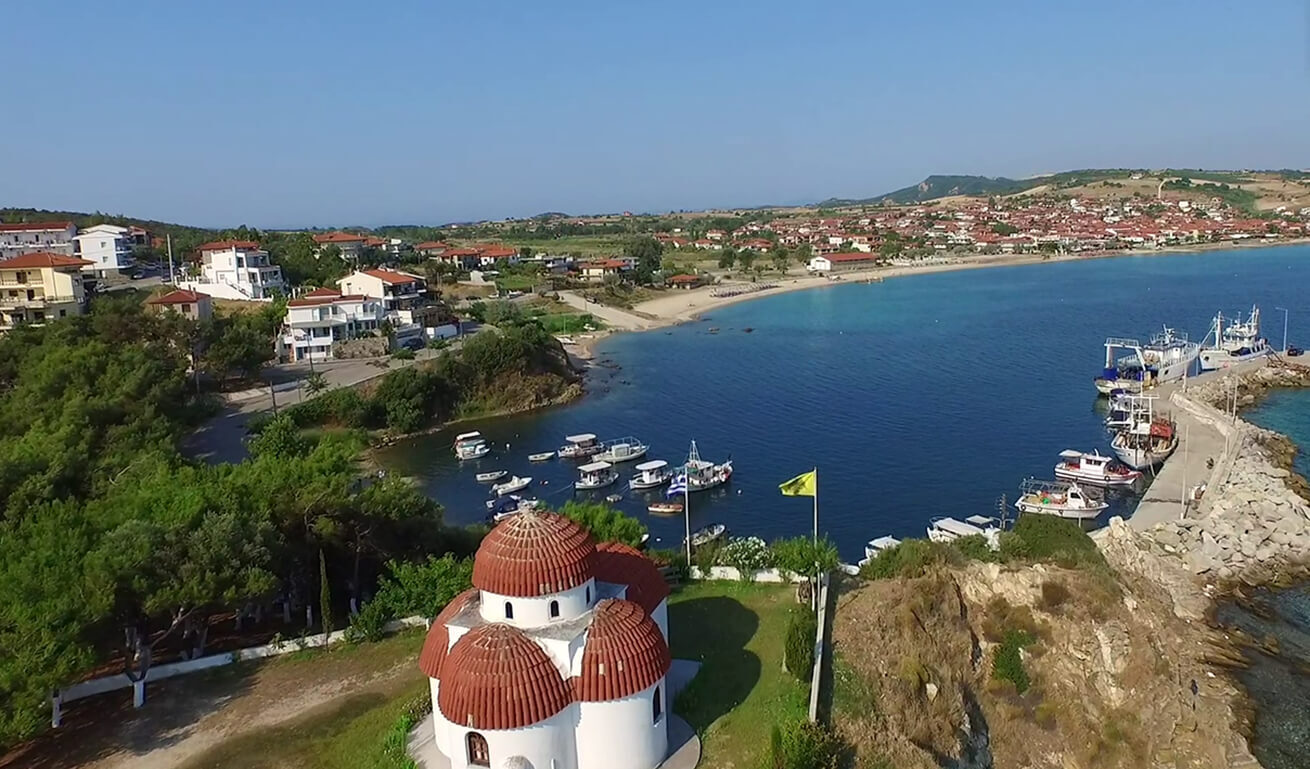 Aerial view of a coastal village with a small church featuring a red dome, several moored boats, and nearby beach.
