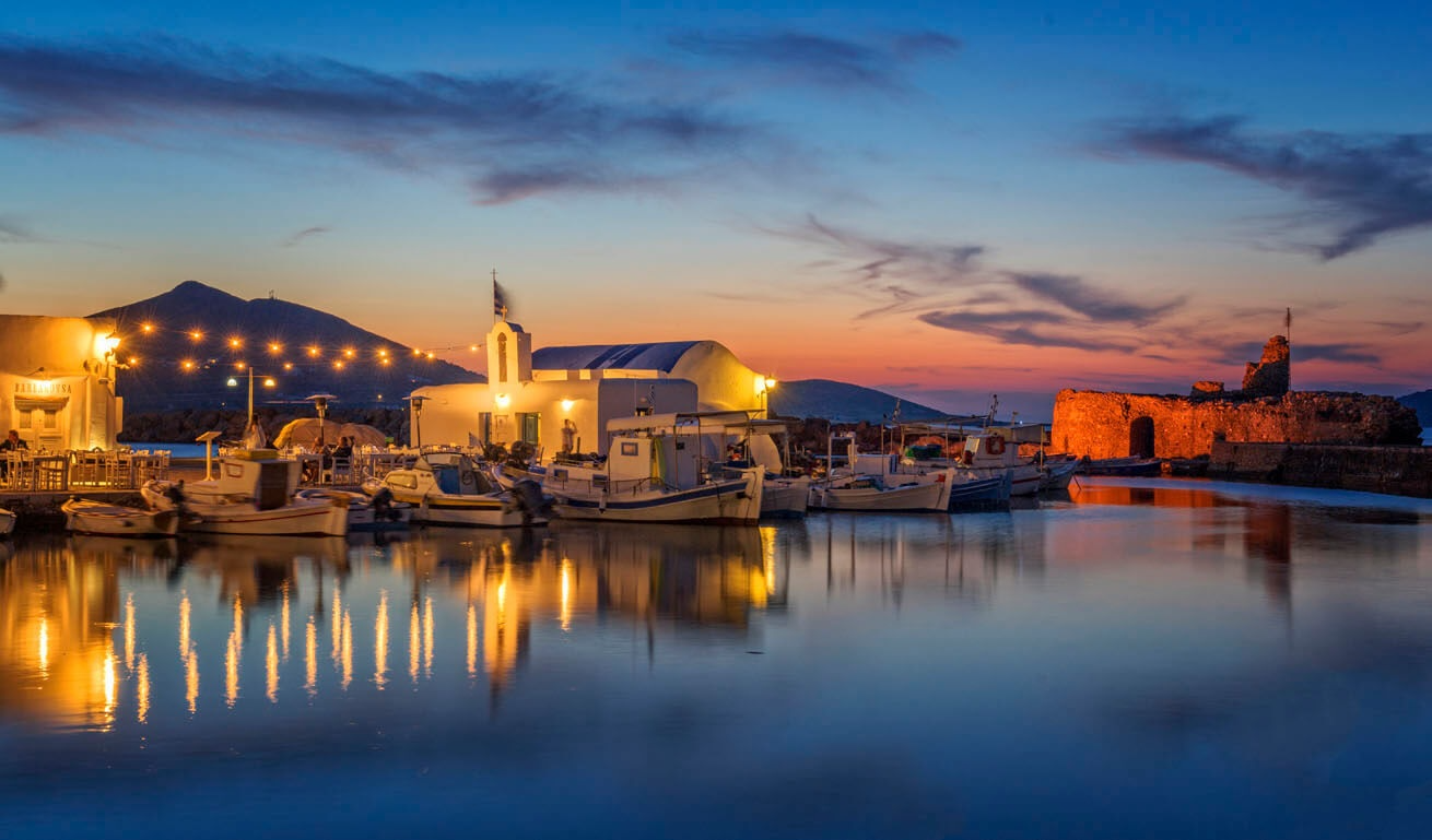 Ήρεμο λιμανάκι με βάρκες και μια μικρή άσπρη εκκλησιά με φώτα το δειλινό.
