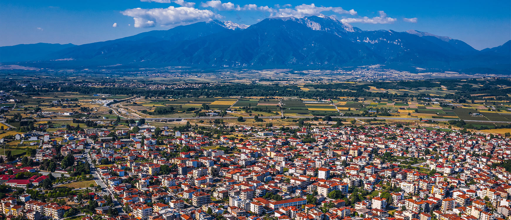 Aerial view of city with low buildings with red roofs. In the background fields and in the distance high mountains. Sunshine.