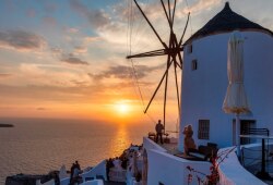 Ηλιοβασίλεμα στην Οία της Σαντορίνης, με παραδοσιακό λευκό ανεμόμυλο να δεσπόζει πάνω από το Αιγαίο Πέλαγος. Άνθρωποι απολαμβάνουν τη θέα από τις βεράντες, ενώ ο ουρανός γεμίζει με ζεστές αποχρώσεις του πορτοκαλί και του ροζ.