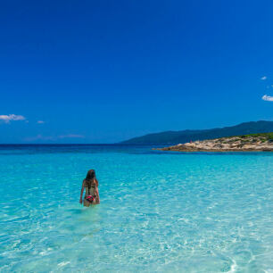 Ρηχή γαλάζια θάλασσα και μια κοπέλα με μαγιό μπαίνει μέσα. Στο βάθος στεριά με χαμηλή βλάστηση.
