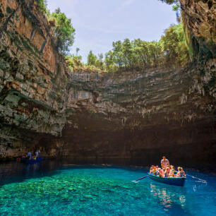 Σπήλαιο με γαλαζοπράσινα νερά και άνοιγμα στην κορυφή που φαίνεται ο ουρανός και δέντρα. 2 βάρκες με ανθρώπους.