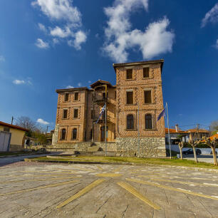 Traditional stone three-story building with closed windows and a tiled roof in front of an open paved area with the symbol of the sun of Vergina. Sunshine.