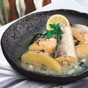 A plate of fish with lemon slices and herbs, accompanied by a glass of white wine, placed on a table with a striped cloth next to a wooden chair.