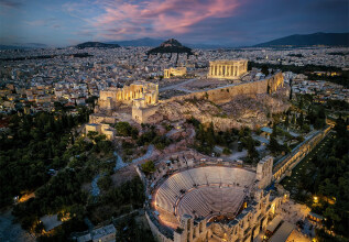 Αεροφωτογραφία της Αθήνας το σούρουπο, με την φωταγωγημένη Ακρόπολη και τον Παρθενώνα πάνω στον λόφο, το εκτεταμένο αστικό τοπίο στο φόντο και τον Λυκαβηττό να διακρίνεται στην απόσταση.