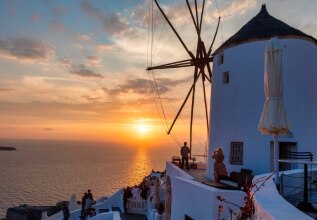 Ηλιοβασίλεμα στην Οία της Σαντορίνης, με παραδοσιακό λευκό ανεμόμυλο να δεσπόζει πάνω από το Αιγαίο Πέλαγος. Άνθρωποι απολαμβάνουν τη θέα από τις βεράντες, ενώ ο ουρανός γεμίζει με ζεστές αποχρώσεις του πορτοκαλί και του ροζ.