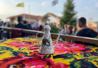 A traditional metal coffee pot sits on a brightly patterned tablecloth at an outdoor setting with blurred people and Greek flags in the background.