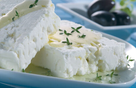 Two pieces of feta cheese with a little oil and fresh oregano on a white plate and a few black olives on a smaller plate. 